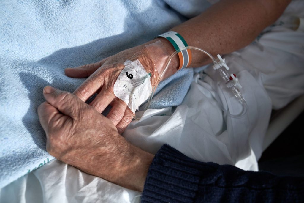 An unseen elderly couple is holding hands. It is implied that the man is sick, as he is receiving an IV infusion, and is being cared for by what appears to be an elderly woman's hand.