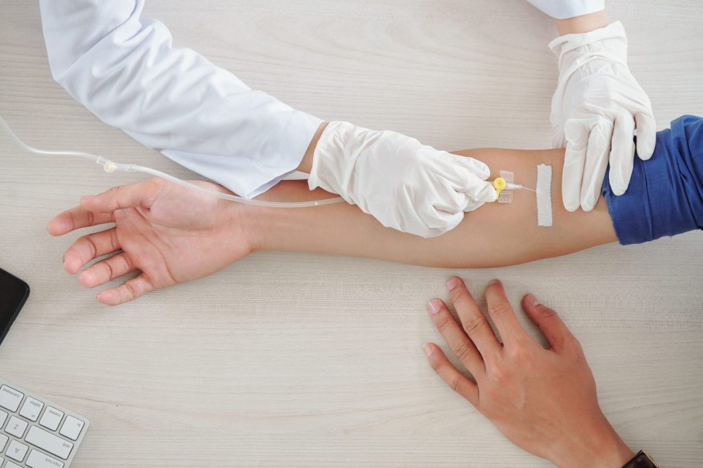 A patient appears to be receiving a type of infusion in their home, with white gloves delivering the infusion into the other person's arm.