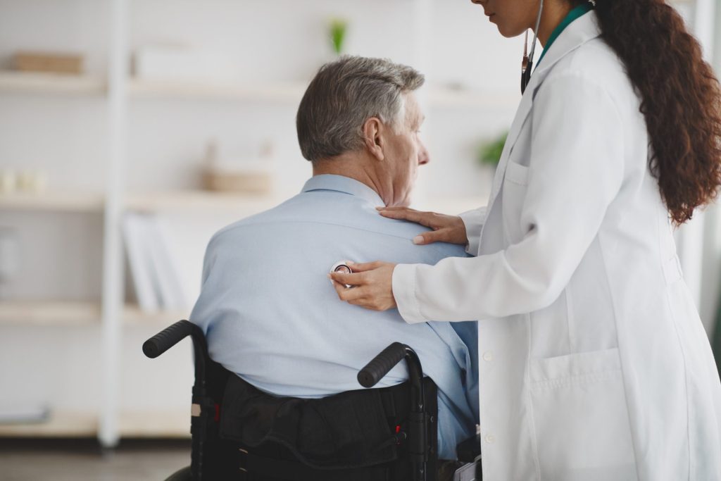 Female doctor is using a stethoscope on an older man who is in a wheelchair.