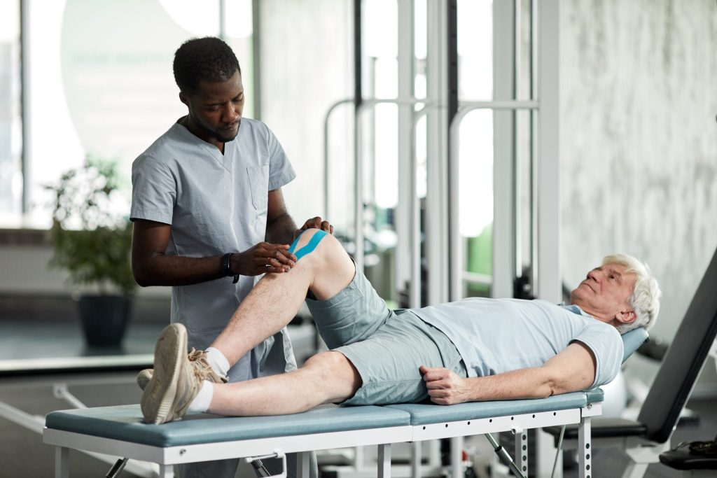 Younger man is performing physical therapy on the knee of an older man that is lying down on a medical table.