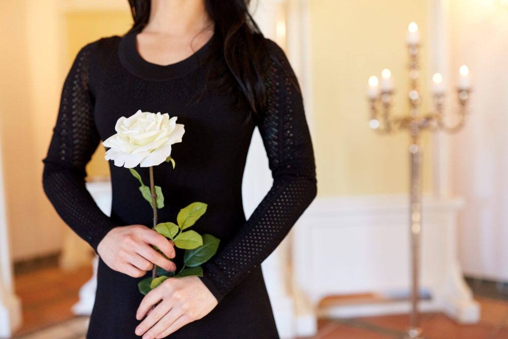 Torso of a woman wearing a black dress in a funeral home, and holding a white flower.