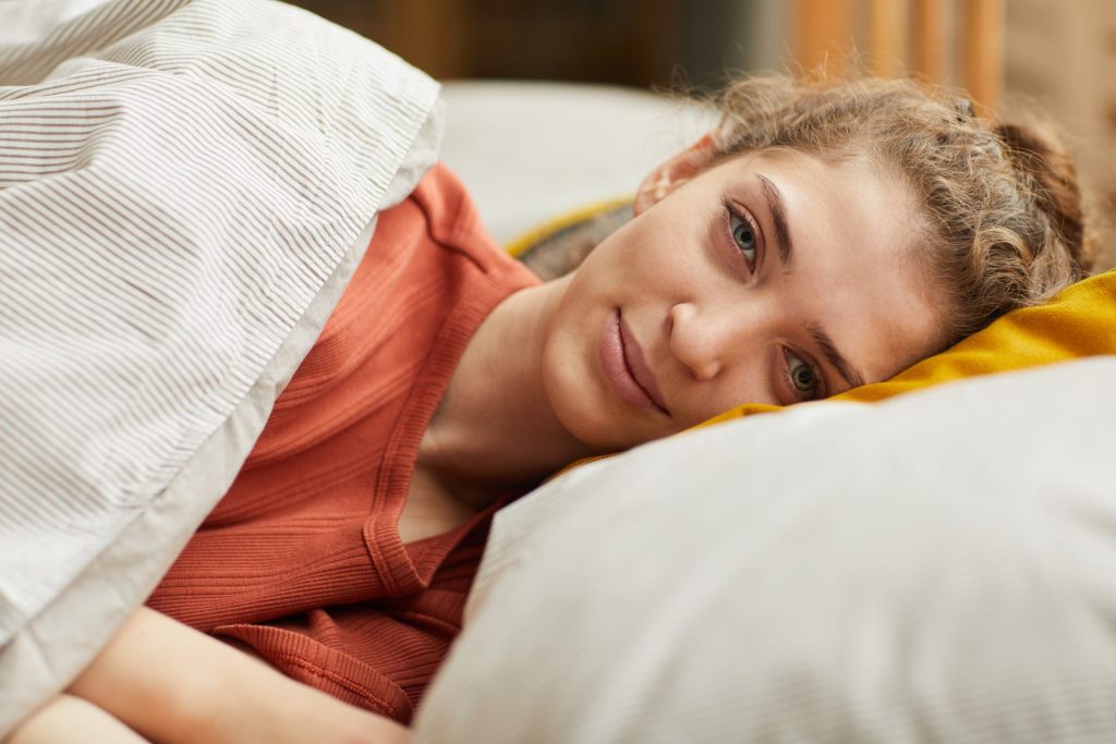 Young woman is laying on a pillow on a bed, looking at the viewer and smiling. She has curly brown hair and is awake, but looks like she experienced a useful rest.